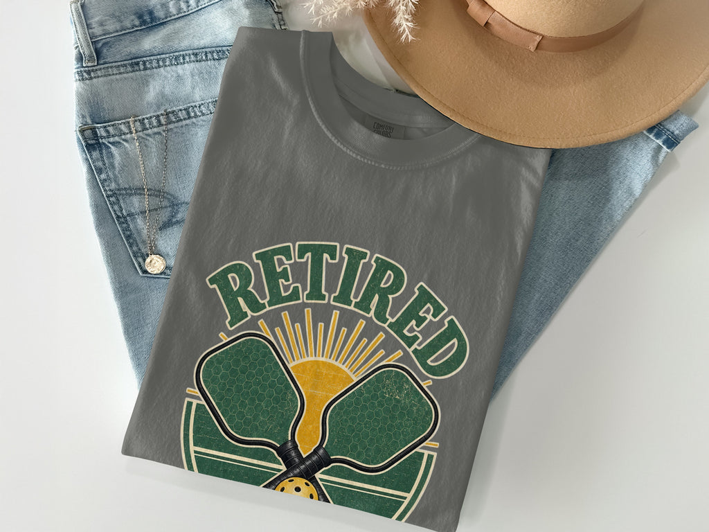Grey t-shirt with green text reading "Retired", pickleball paddles, and a sun graphic.