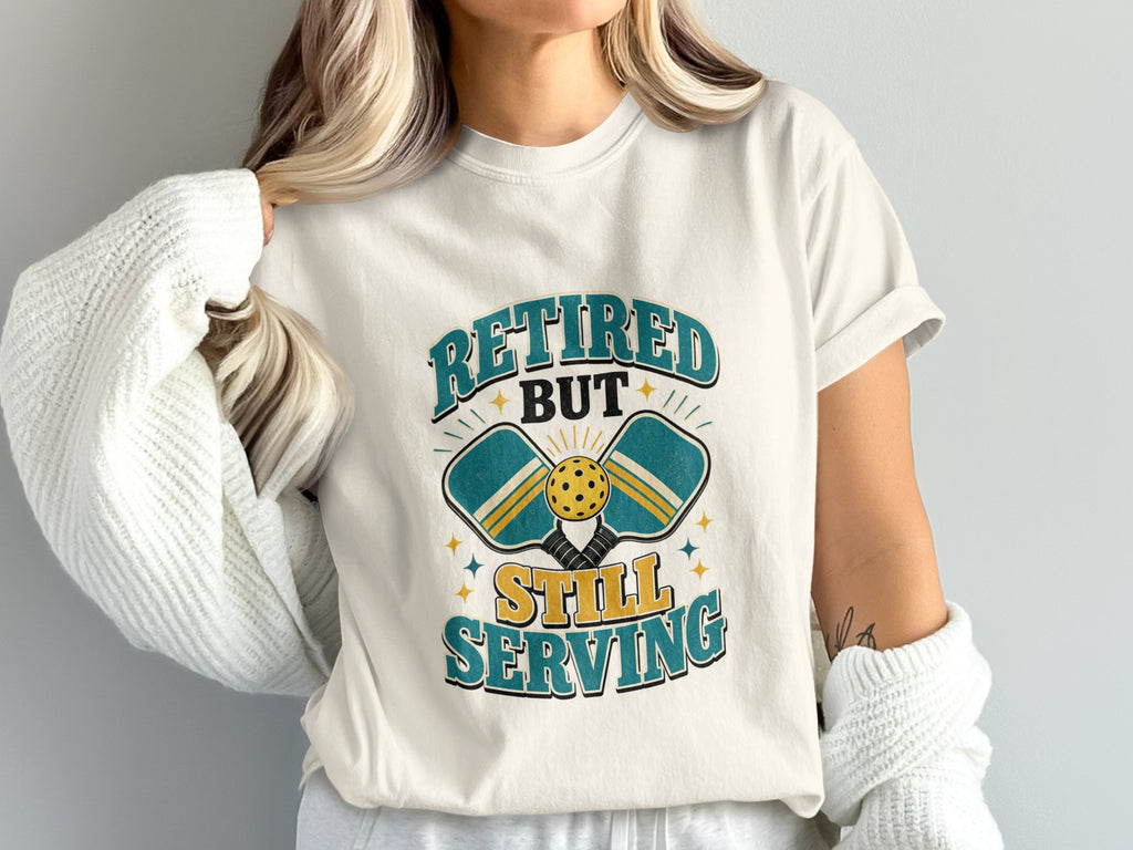 T-shirt graphic featuring pickleball paddles, ball, and text: "Retired But Still Serving".
