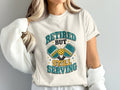 T-shirt graphic featuring pickleball paddles, ball, and text: "Retired But Still Serving".