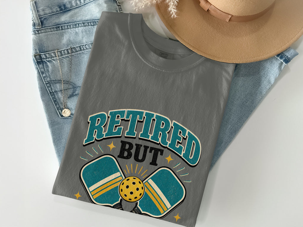 Grey t-shirt with "Retired But" text and pickleball paddles.